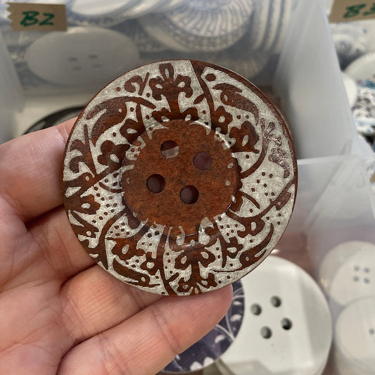 Hand holding a decorative button with intricate design over a container of buttons.