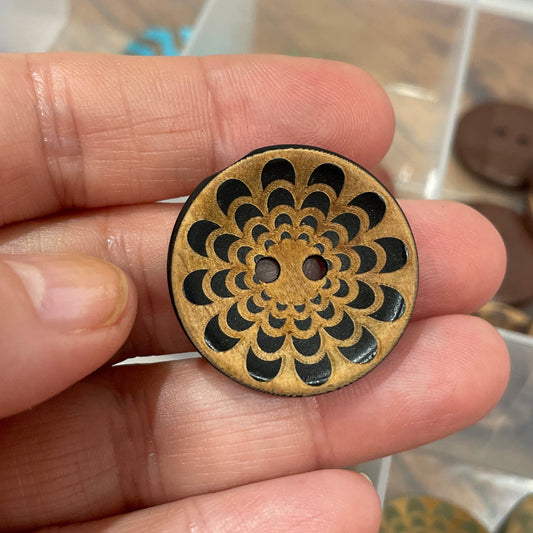 Hand holding a decorative wooden button with intricate design, against a background of similar buttons in a container.