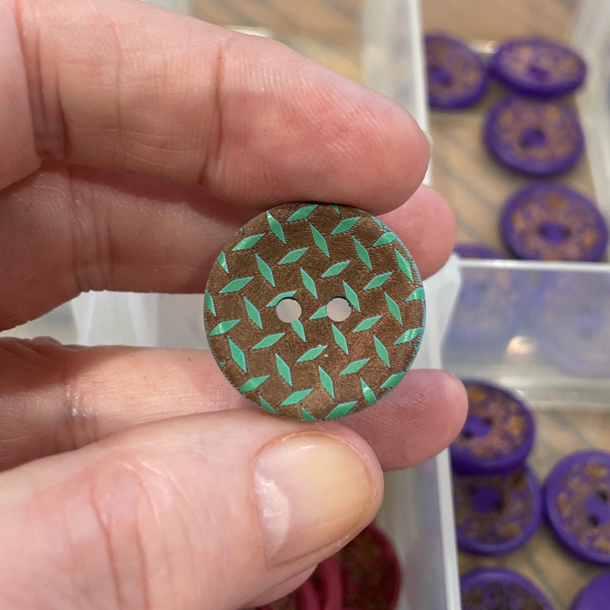 Hand holding a green button with a pattern, with a container of buttons in the background.