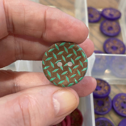 Hand holding a green button with a pattern, with a container of buttons in the background.