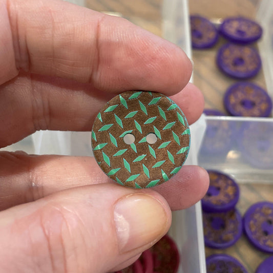 Hand holding a green button with a pattern, with a container of buttons in the background.