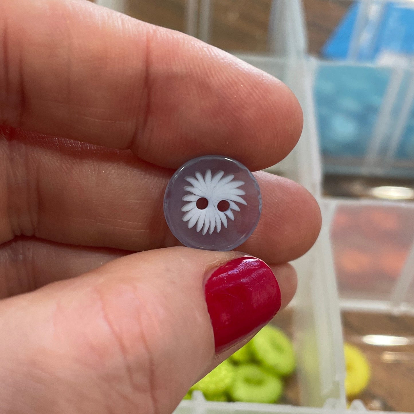 Hand holding a small button with a decorative center, against a background of a container with various buttons.