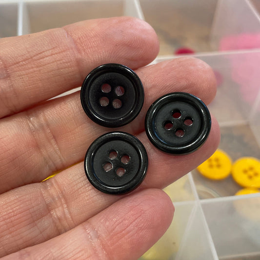 Three black buttons held in a hand with a container of colorful buttons in the background.