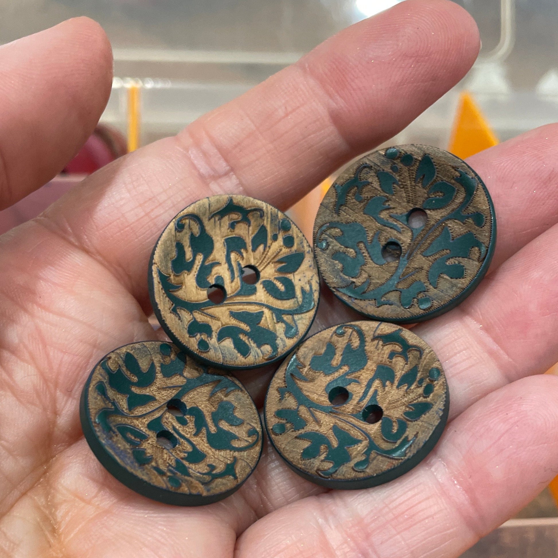Hand holding four decorative buttons with intricate designs, with a blurred background of more buttons.