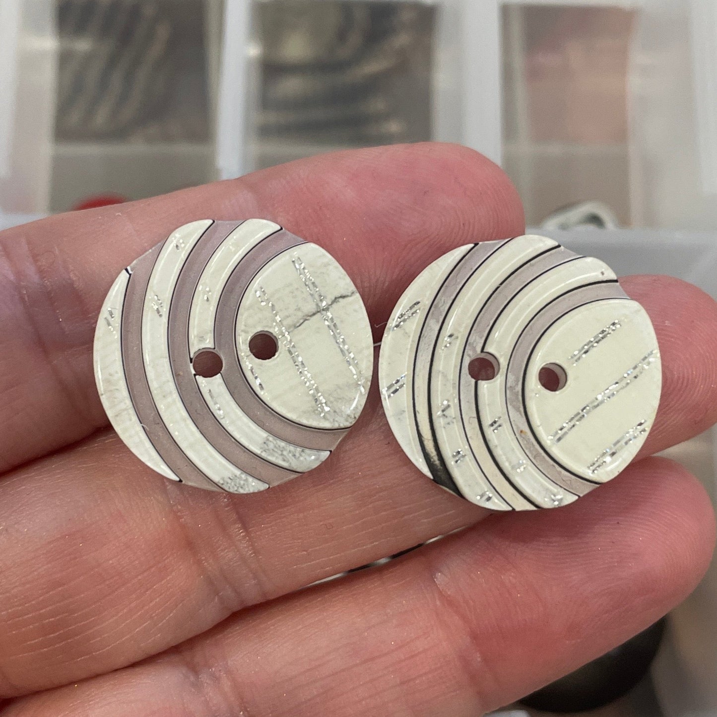 Hand holding two white buttons with brown accents in front of a container of buttons.