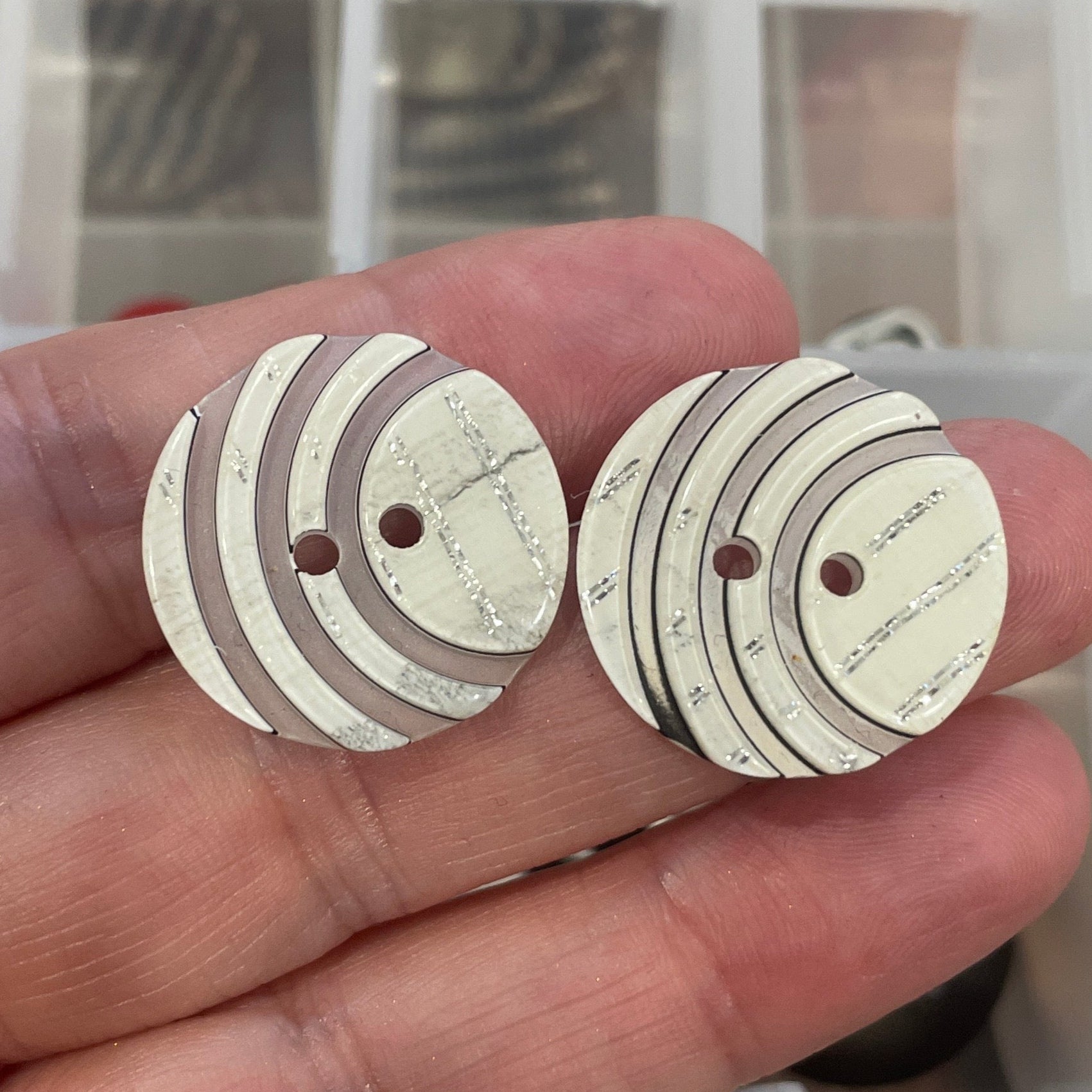 Hand holding two white buttons with brown accents in front of a container of buttons.