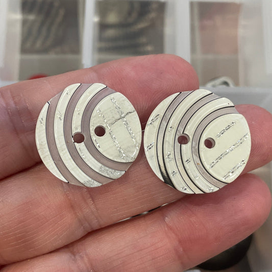Hand holding two white buttons with brown accents in front of a container of buttons.
