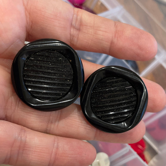 Black textured buttons held in a hand with a container of embellishments in the background.