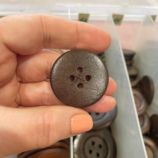 Hand holding a dark brown button with four holes, with a collection of similar buttons in the background.