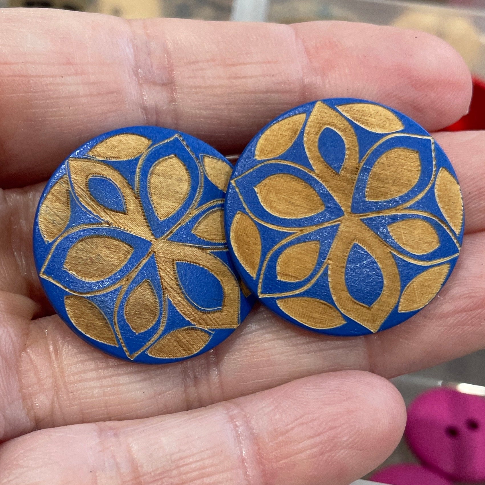 Hand holding two decorative buttons with a blue and gold pattern, with a display of more buttons in the background.