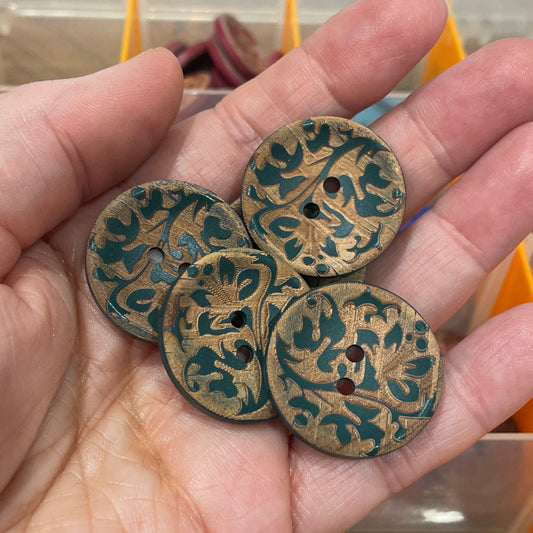 Hand holding three decorative buttons with intricate patterns in front of a container filled with various buttons.