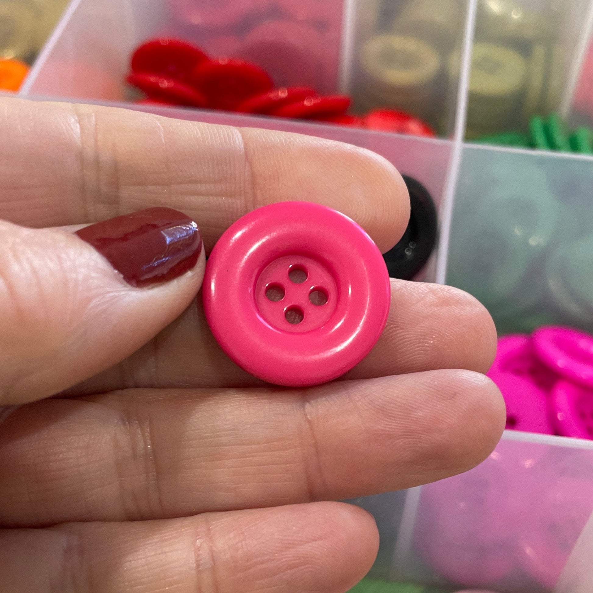 Hand holding a pink button with a container of colorful buttons in the background