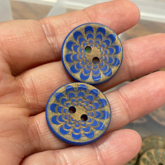 Hand holding two blue and gold decorative buttons with a container of similar buttons in the background.