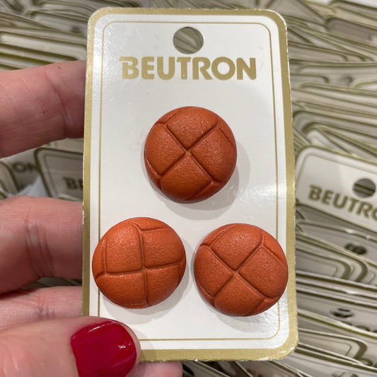 Three brown textured buttons on a 'Beutron' card held by a hand with red nail polish.