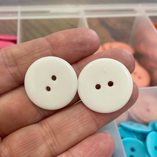 Two white buttons held in a hand with a container of colorful buttons in the background.