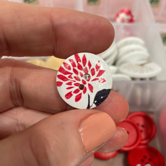 Hand holding a button with a red floral design, surrounded by various buttons in the background.