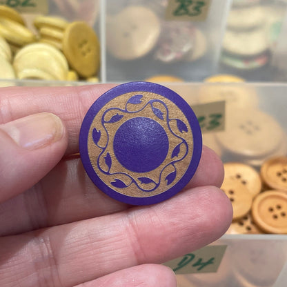 Hand holding a decorative button with a purple center, surrounded by various buttons in a clear container.