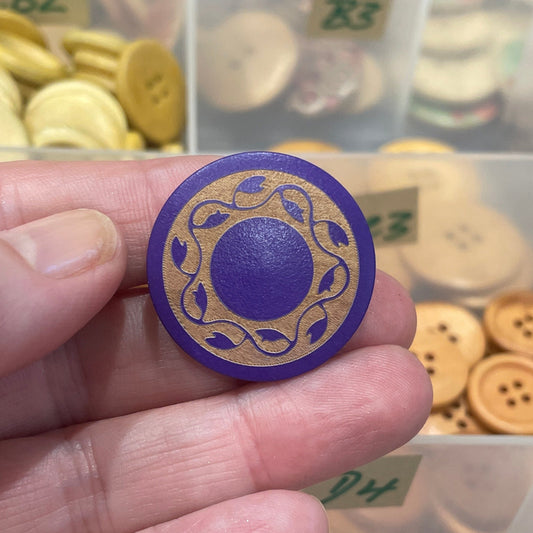 Hand holding a decorative button with a purple center, surrounded by various buttons in a clear container.