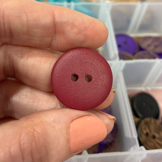 Maroon button held between fingers with a container of buttons in the background