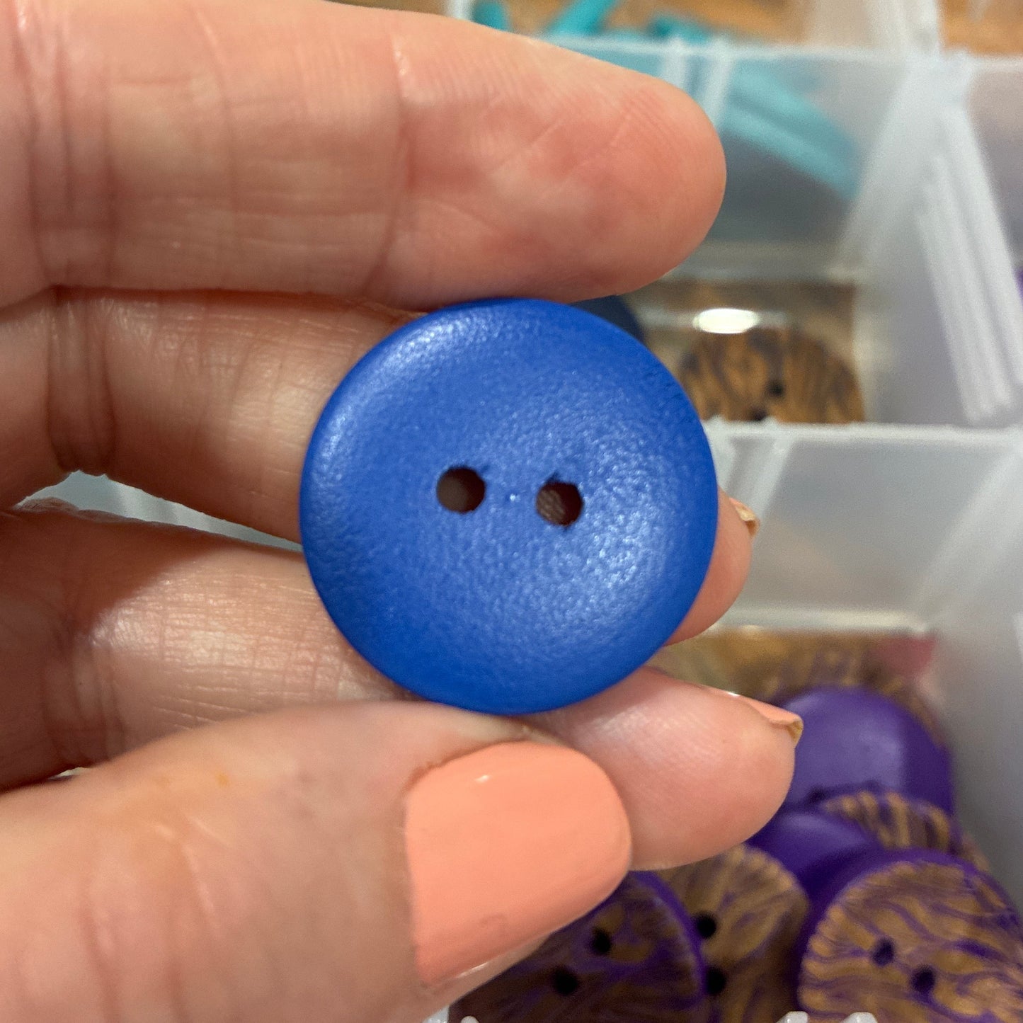 Hand holding a blue button with a blurred background of a storage box with buttons.