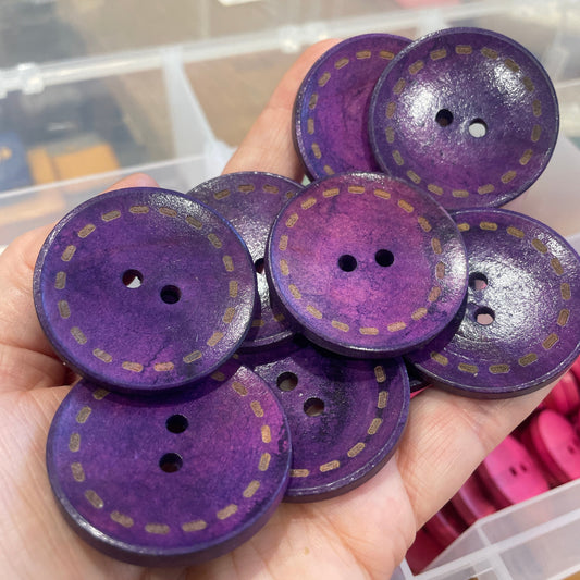 Purple buttons with gold stitching held in a hand against a blurred background