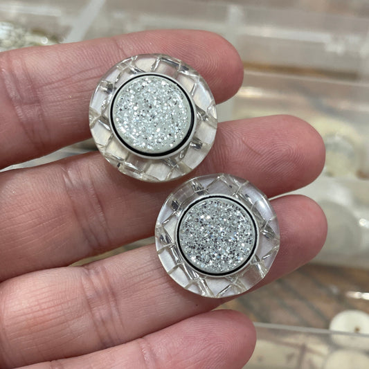 Silver glittery buttons held between fingers with a blurred background of red and clear containers.