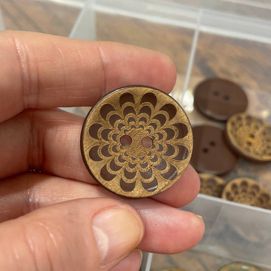 Hand holding a decorative wooden button with a detailed design, against a background of similar buttons in a box.
