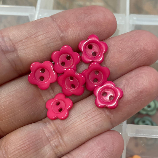 Hand holding pink flower-shaped buttons with a container of similar buttons in the background.