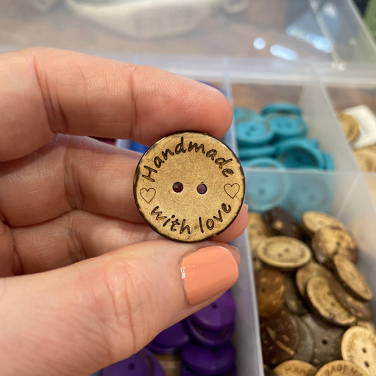 button with 'Handmade with Love' text held by a hand, with a container of buttons in the background.