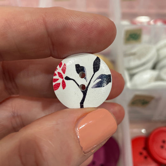 Hand holding a decorative button with a floral design in front of a container of various buttons.