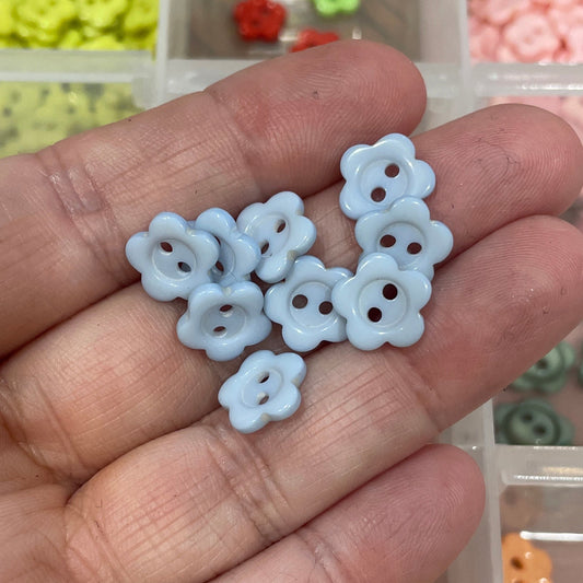Hand holding small white flower-shaped beads with a container of assorted beads in the background.