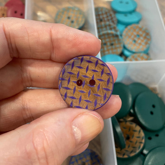Hand holding a purple button with a pattern, with a container of buttons in the background.