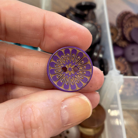 Hand holding a purple button with a decorative center, against a background of various buttons in a container.