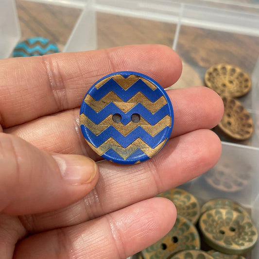 Hand holding a blue and gold zigzag patterned button with a collection of buttons in the background.