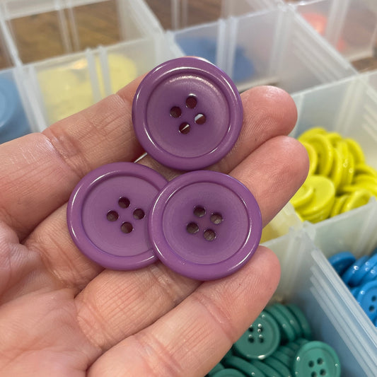 Hand holding three purple buttons with a container of colorful buttons in the background.