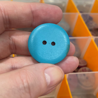 Hand holding a blue button with a clear container of buttons in the background