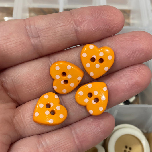 Hand holding orange heart-shaped buttons with white polka dots, with a container of buttons in the background.