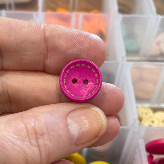 Hand holding a pink button with a container of colorful buttons in the background.