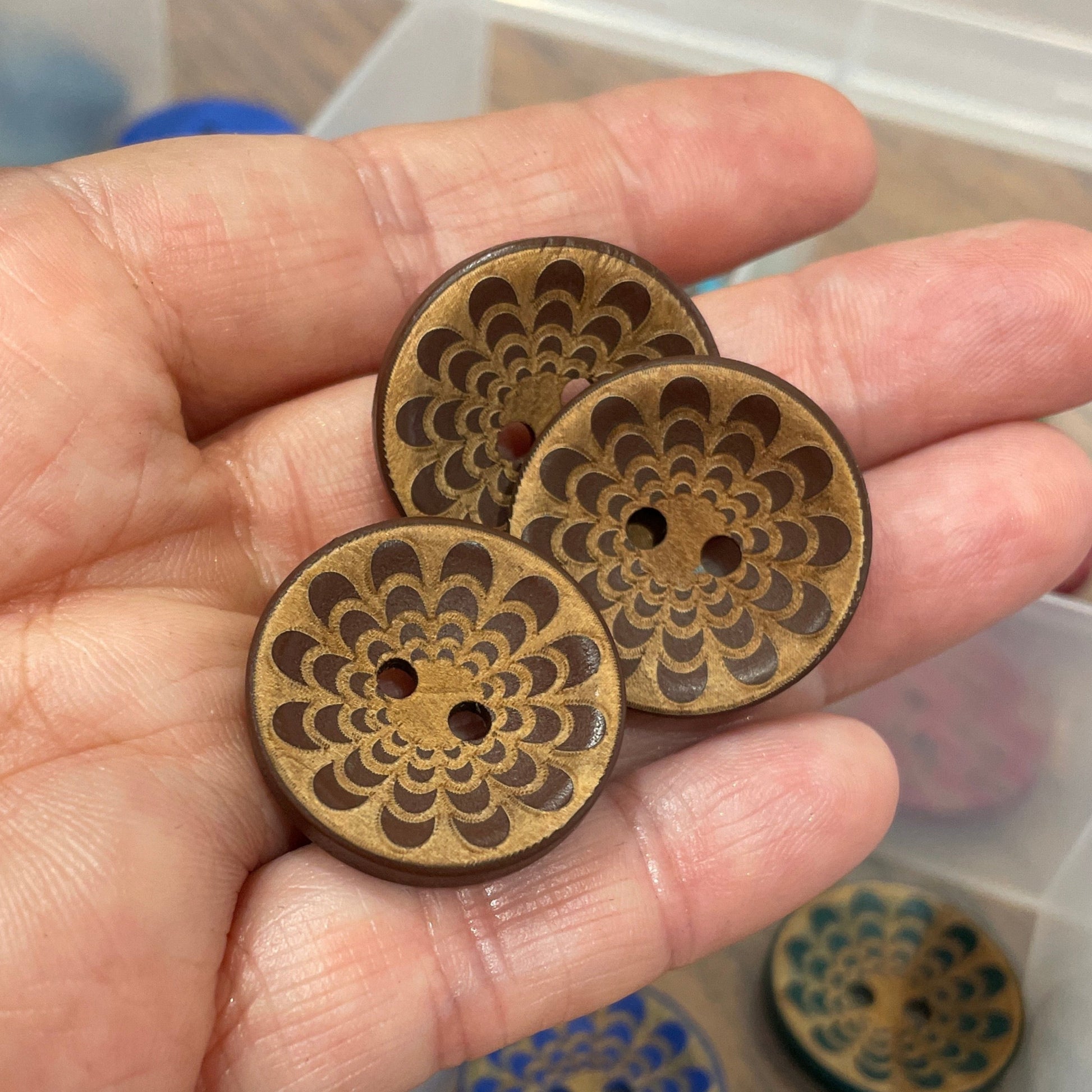 Hand holding three wooden buttons with intricate designs, with a clear plastic organizer in the background.
