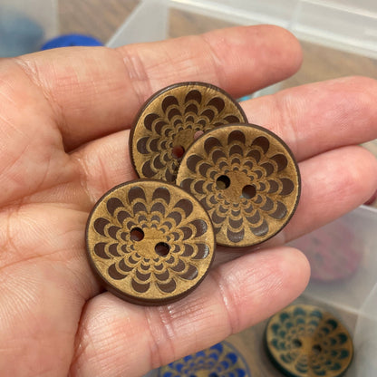 Hand holding three wooden buttons with intricate designs, with a clear plastic organizer in the background.