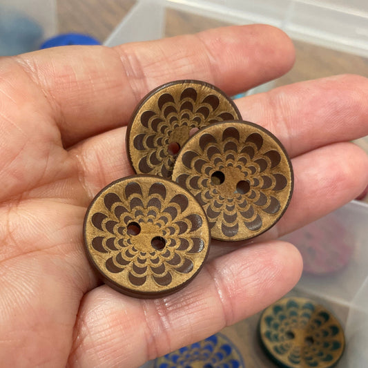 Hand holding three wooden buttons with intricate designs, with a clear plastic organizer in the background.