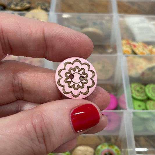 Hand holding a pink floral button with a clear jewelry box containing various button in the background.