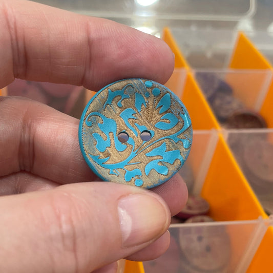 Hand holding a decorative button with a background of a compartmentalized container filled with various buttons.