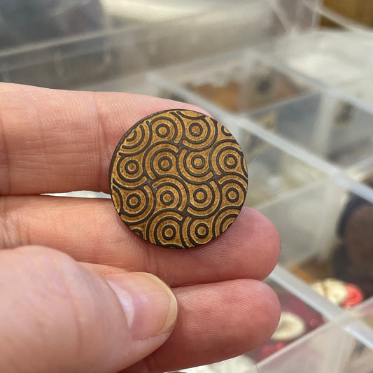 Hand holding a button with intricate patterns in front of a clear container with various items.
