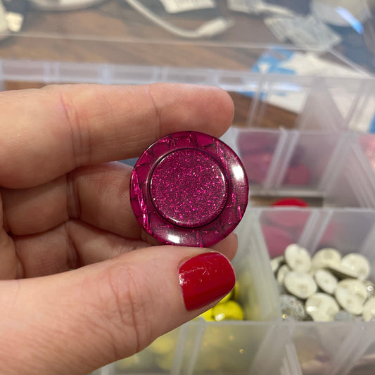 Hand holding a pink glittery button with a container of buttons in the background