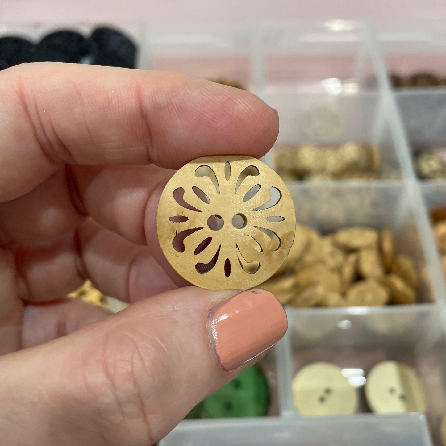 Hand holding a gold decorative button with a detailed design, against a blurred background of a button collection.