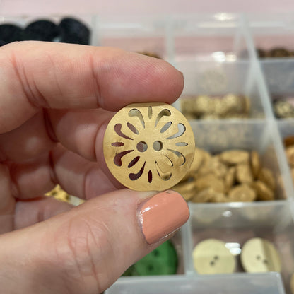Hand holding a gold decorative button with a detailed design, against a blurred background of a button collection.
