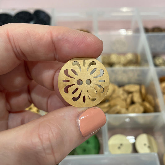 Hand holding a gold decorative button with a detailed design, against a blurred background of a button collection.