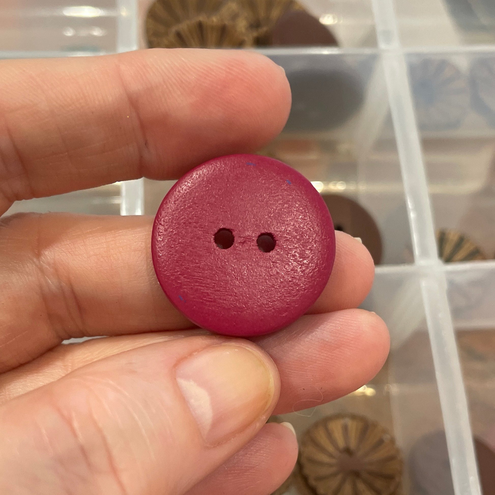 Hand holding a pink button with a background of assorted buttons in a clear container.