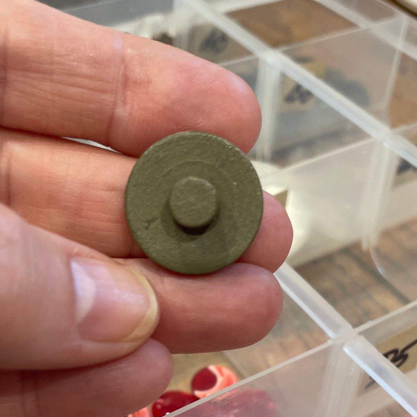 Hand holding a small round button with a background of a button collection in a clear organizer.